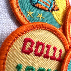May include: Embroidered patches with orange, yellow, and teal accents. One patch features a rocket ship design, while another displays the word "DOLL" in red and "100" in green. The patches have an orange border.