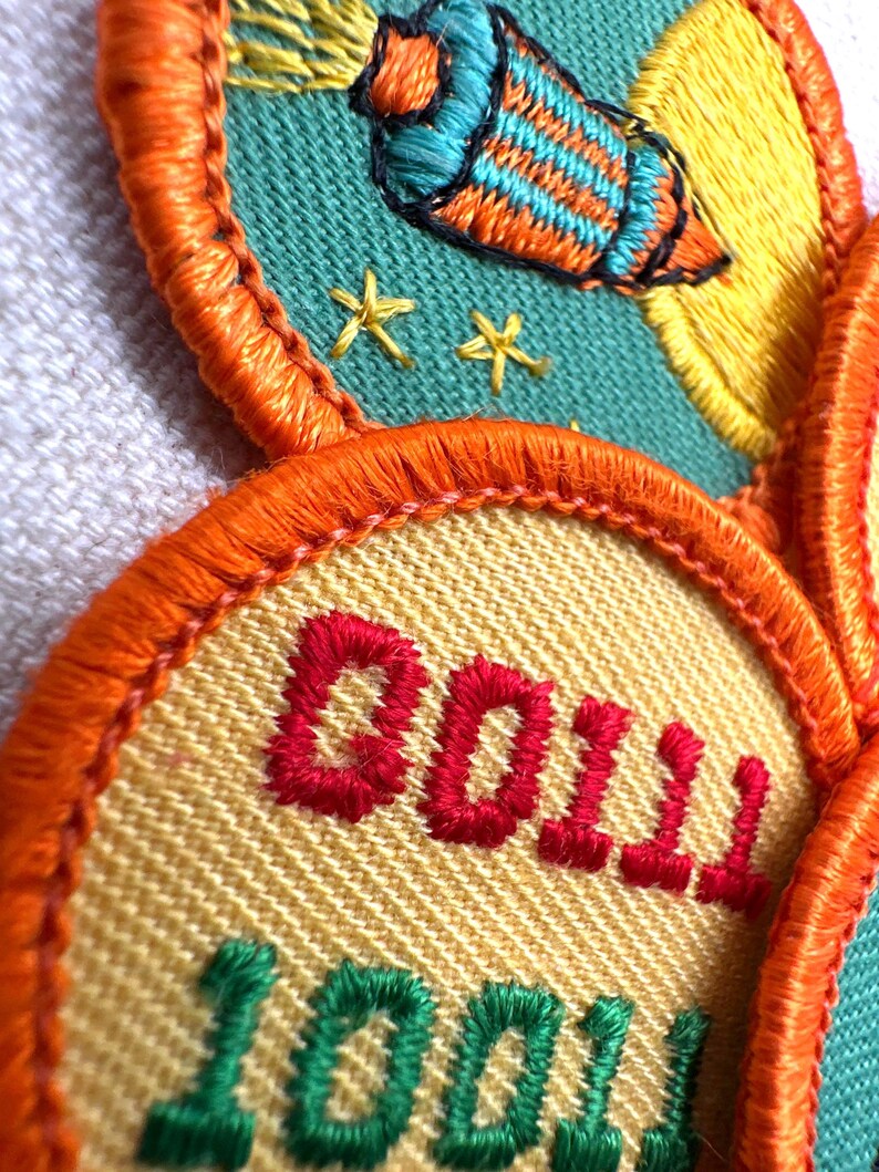 May include: Embroidered patches with orange, yellow, and teal accents. One patch features a rocket ship design, while another displays the word "DOLL" in red and "100" in green. The patches have an orange border.