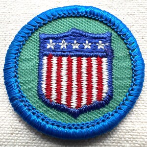 May include: A round patch with a blue border and a green interior. The patch features a shield design with red and white stripes and a blue top with three white stars. The patch is likely an emblem or insignia.
