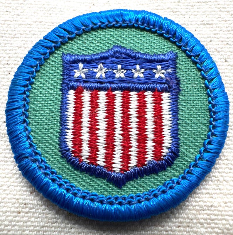 May include: A round patch with a blue border and a green interior. The patch features a shield design with red and white stripes and a blue top with three white stars. The patch is likely an emblem or insignia.