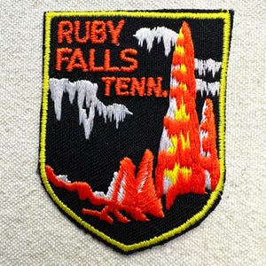 May include: Embroidered patch with the words "RUBY FALLS TENN." in orange. The patch features a black background with white and orange mountain-like shapes and a yellow border.