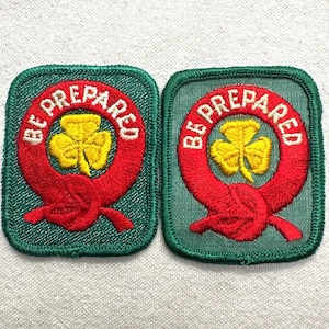 May include: Two green embroidered patches with red and yellow details. Each patch features a red circular design with the words "BE PREPARED" and a yellow shamrock in the center. The patches are square with rounded corners.