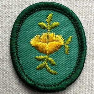May include: An oval-shaped embroidered patch featuring a vibrant yellow flower with green leaves centered on a solid green background. The patch is finished with a dark green stitched border that frames the entire design in a clean, circular edge.