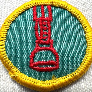 May include: Round patch with a green center and a red embroidered design. The design appears to be a stylized torch or flame. The patch is bordered with yellow stitching.