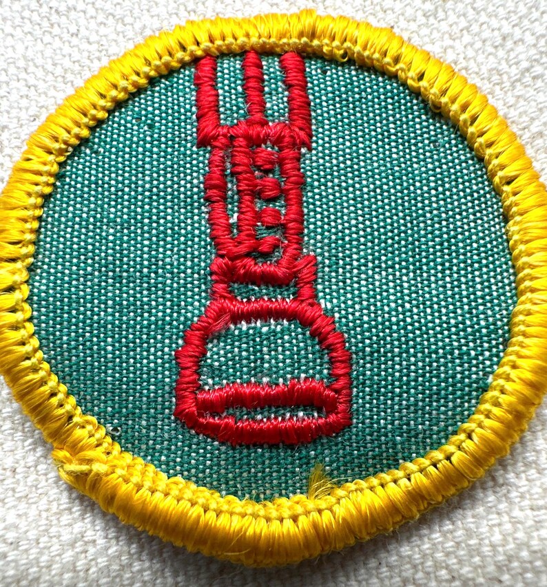 May include: Round patch with a green center and a red embroidered design. The design appears to be a stylized torch or flame. The patch is bordered with yellow stitching.