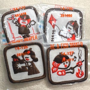 May include: Four square embroidered patches with brown borders and white backgrounds. Each patch features a cartoon figure in brown and red, with text and numbers. The patches are packaged in clear plastic.