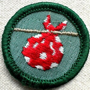May include: Embroidered patch featuring a red and white dotted bag suspended from a tan line. The bag is set against a teal background and framed by a green stitched border. A classic design for a scout or outdoor enthusiast.