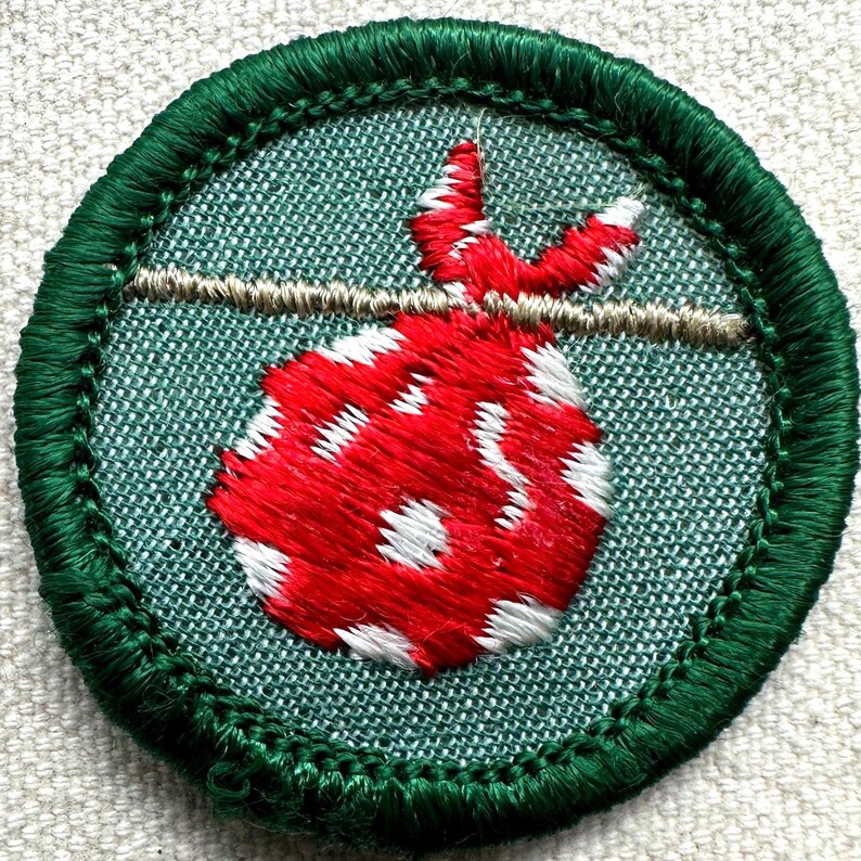 May include: Embroidered patch featuring a red and white dotted bag suspended from a tan line. The bag is set against a teal background and framed by a green stitched border. A classic design for a scout or outdoor enthusiast.