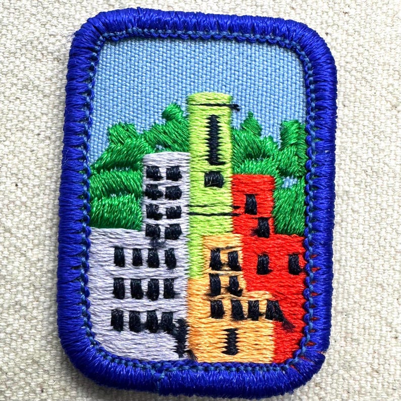May include: Embroidered patch featuring a colorful cityscape with buildings in shades of gray, green, yellow, and red. The patch has a blue border and a light blue sky background, depicting a vibrant urban scene.