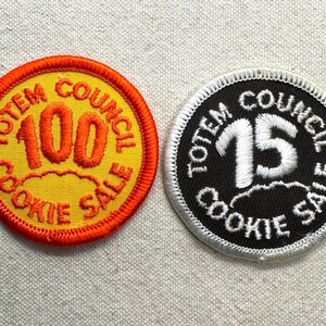 May include: Two circular embroidered patches. The left patch is yellow with an orange border and features the orange text "TOTEM COUNCIL COOKIE SALE" surrounding the number "100". The right patch is black with a white border and white text.