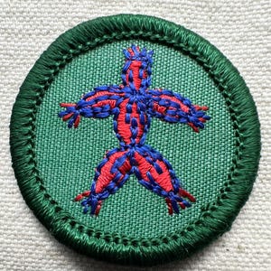 May include: Embroidered patch featuring a stylized figure with outstretched arms and legs. The figure is red with blue outlining and details, set against a green fabric background. The patch has a green border.