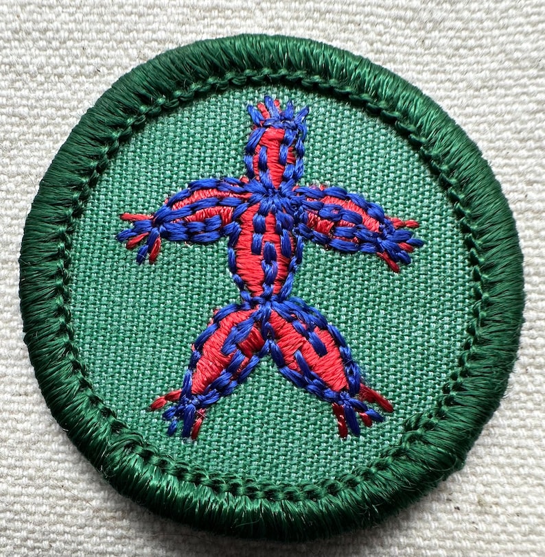 May include: Embroidered patch featuring a stylized figure with outstretched arms and legs. The figure is red with blue outlining and details, set against a green fabric background. The patch has a green border.
