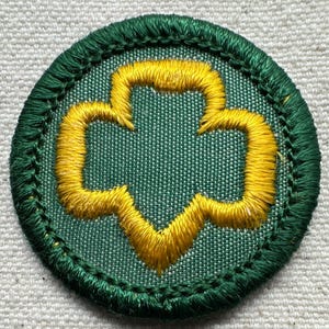 May include: A round, embroidered patch with a green border and a green background. The patch features a yellow trefoil design in the center. The patch is likely a Girl Scout badge.