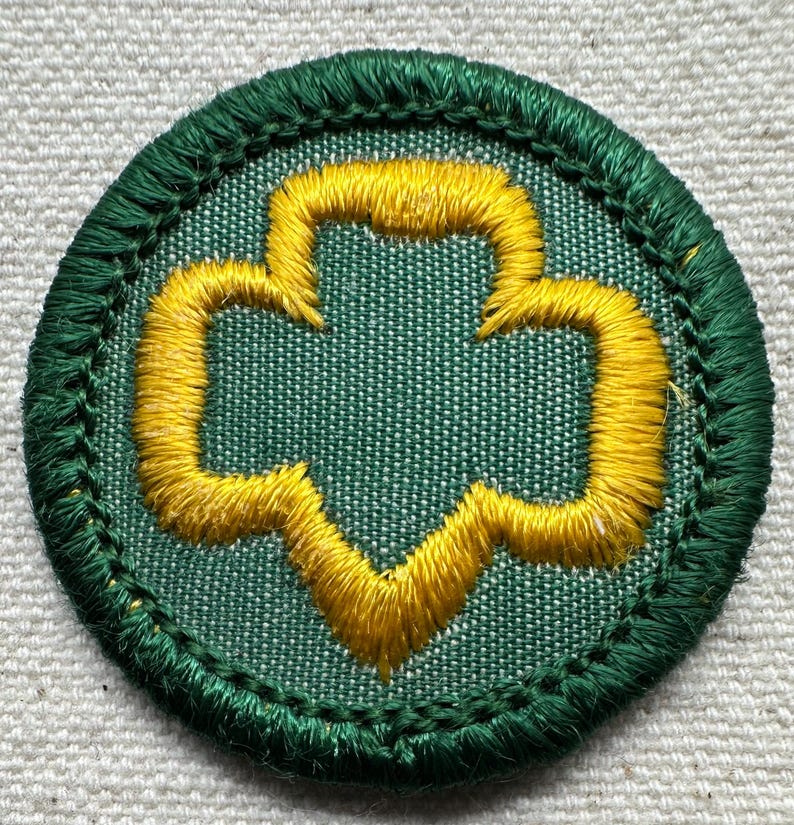 May include: A round, embroidered patch with a green border and a green background. The patch features a yellow trefoil design in the center. The patch is likely a Girl Scout badge.