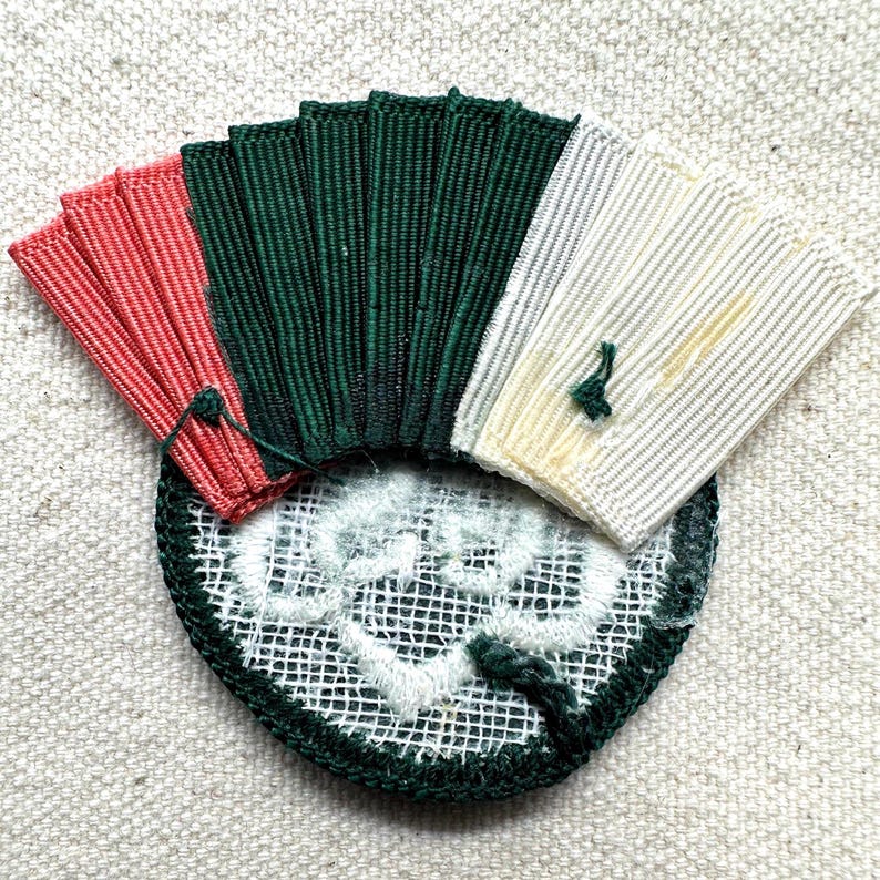 May include: A detailed, handmade textile patch with a fan-like design. The patch features sections of red, dark green, and cream-colored fabric, all attached to a circular base with a white and green pattern. The patch is likely for decorative purposes.