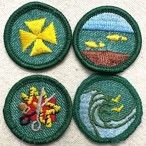 May include: Four round embroidered patches with green borders. Designs include a yellow cross, a landscape with water and fish, a floral design with scissors, and a wave with birds. The patches are on a textured, light background.