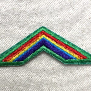 Puede incluir: Parche bordado con dise&ntilde;o de gal&oacute;n en colores del arco&iacute;ris: rojo, naranja, amarillo, verde, azul y morado. El parche tiene un borde verde y est&aacute; sobre un fondo texturizado blanquecino. Ideal para chaquetas, bolsos u otros art&iacute;culos de tela.
