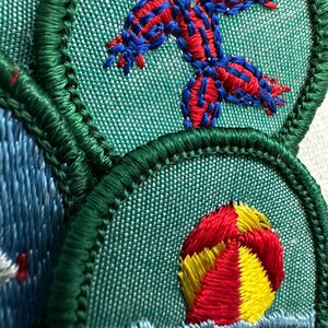 Puede incluir: Primer plano de parches bordados con un borde verde. Un parche presenta un dise&ntilde;o de estrella de mar roja y azul sobre un fondo verde. Otro parche muestra una pelota de playa roja, amarilla y blanca sobre agua azul. Los parches son probablemente para una organizaci&oacute;n de exploradores o juvenil.
