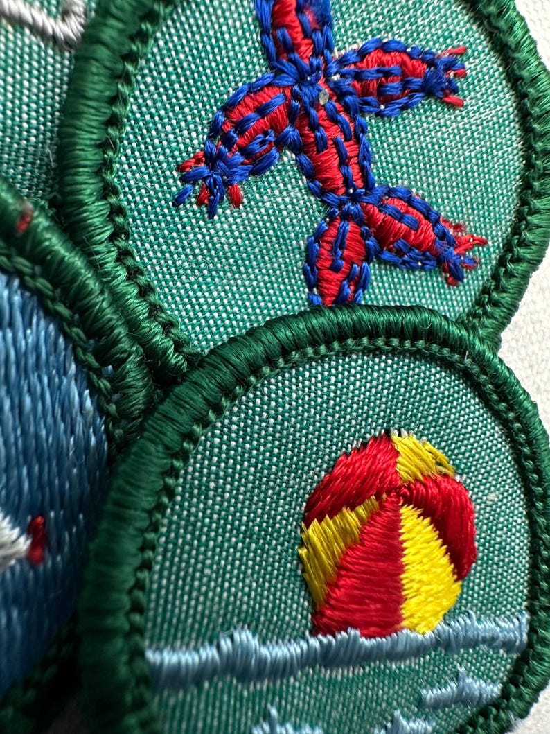Puede incluir: Primer plano de parches bordados con un borde verde. Un parche presenta un dise&ntilde;o de estrella de mar roja y azul sobre un fondo verde. Otro parche muestra una pelota de playa roja, amarilla y blanca sobre agua azul. Los parches son probablemente para una organizaci&oacute;n de exploradores o juvenil.