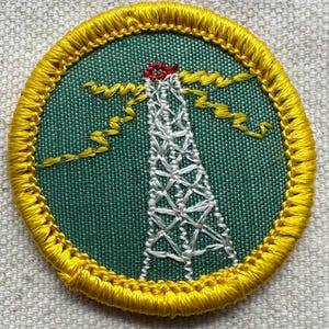 May include: Embroidered patch featuring a white radio tower with a red top, set against a green background. Yellow zigzag lines emanate from the tower, and the patch is bordered by a yellow stitched edge. The patch is circular.