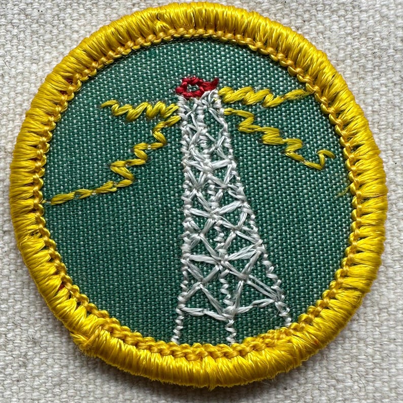 May include: Embroidered patch featuring a white radio tower with a red top, set against a green background. Yellow zigzag lines emanate from the tower, and the patch is bordered by a yellow stitched edge. The patch is circular.
