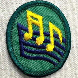 May include: An embroidered patch with a green oval border. The patch features yellow musical notes over blue horizontal lines, set against a green background. The design is a symbol of music.