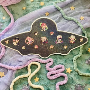 May include: A black, glitter-covered, UFO-shaped tray with cartoon character pins. The tray is surrounded by light purple, green, and white yarn, gold star-shaped decorations, and a green and blue textured fabric.