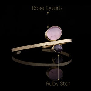 May include: A gold-toned bracelet featuring a Rose Quartz stone and a Ruby Star stone. The bracelet has a textured band and a modern design. The background is black, highlighting the jewelry's reflective surface.