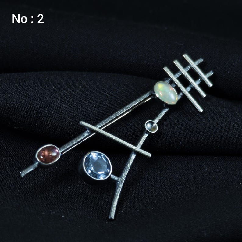 May include: A silver-toned brooch with an abstract design. It features a series of intersecting lines and geometric shapes, adorned with several gemstones in shades of brown, blue, and white. The brooch is set against a dark background, with the text "No: 2" in the upper left corner.