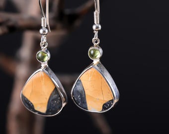 May include: A pair of silver dangle earrings. Each earring features a triangular, orange and black stone set in a silver frame, with a small green stone and a silver hook. The earrings are displayed against a dark background.