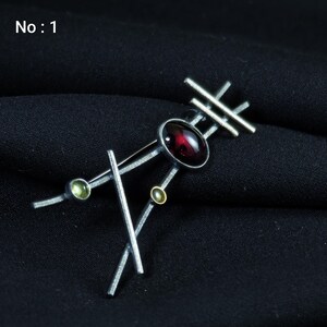 May include: Silver-toned brooch with an abstract design. It features a large, oval, deep red gemstone at the center, accented by smaller green and gold-colored stones. The brooch has a geometric arrangement of straight lines and the text "No: 1".