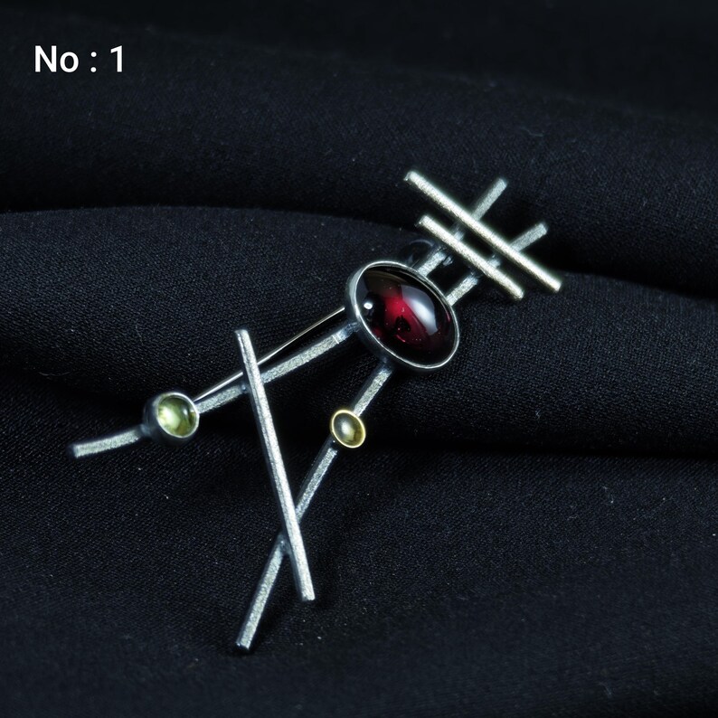 May include: Silver-toned brooch with an abstract design. It features a large, oval, deep red gemstone at the center, accented by smaller green and gold-colored stones. The brooch has a geometric arrangement of straight lines and the text "No: 1".