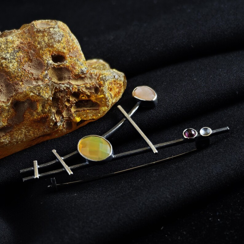 May include: A silver-toned brooch with an abstract design. It features an oval-shaped, yellow-green gemstone, a pink gemstone, and two smaller gemstones. The brooch has a geometric arrangement of straight lines and is set against a black background.