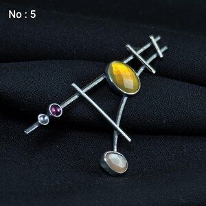May include: Silver-toned brooch with an abstract design. It features a large, oval, yellow gemstone, a smaller, light-colored stone, and two small, round, purple and silver accents. The brooch has a geometric, linear structure.