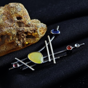 May include: A silver-toned brooch with an abstract design. It features a large, light yellow gemstone, several smaller red and blue stones, and a series of silver bars. The brooch is set against a black background.