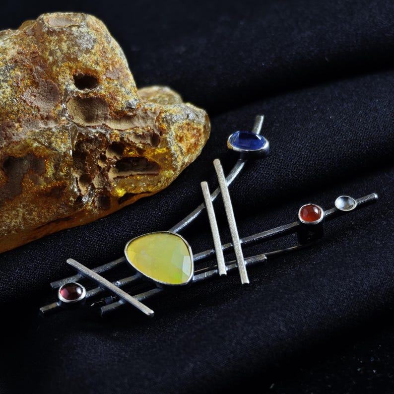 May include: A silver-toned brooch with an abstract design. It features a large, light yellow gemstone, several smaller red and blue stones, and a series of silver bars. The brooch is set against a black background.