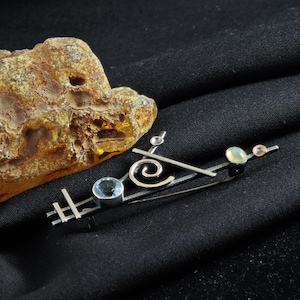 May include: A silver-toned brooch with a geometric design, featuring a blue gemstone, a swirl, and two smaller, round gemstones. The brooch is set against a black background, with a piece of amber in the upper left corner.
