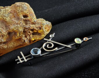 May include: A silver-toned brooch with a geometric design, featuring a blue gemstone, a swirl, and two smaller, round gemstones. The brooch is set against a black background, with a piece of amber in the upper left corner.