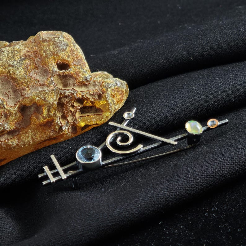 May include: A silver-toned brooch with a geometric design, featuring a blue gemstone, a swirl, and two smaller, round gemstones. The brooch is set against a black background, with a piece of amber in the upper left corner.