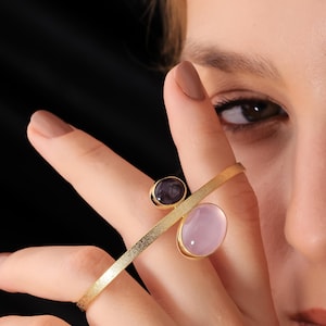 Könnte beinhalten: Ein goldfarbener Ring mit zwei ovalen Edelsteinen, einem dunklen Lila und einem hellen Rosa. Der Ring hat ein modernes, minimalistisches Design mit einem horizontalen Balken. Die Edelsteine sind in goldenen Fassungen gefasst.