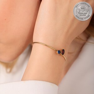 Könnte beinhalten: Ein goldfarbenes Armband mit zwei Edelstein-Akzenten. Einer ist tiefrot, der andere tiefblau. Das Armband ist dünn und filigran, mit einem minimalistischen Design. Das Bild enthält auch ein silbernes Emblem mit der Aufschrift "100% Handmade Sterling Silver Genuine Stone".