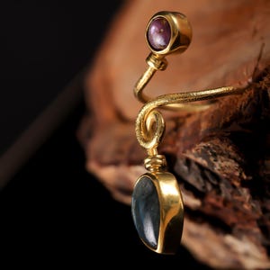 May include: A gold-toned, spiral-shaped ring with a purple gemstone at the top and a dark, oval-shaped stone pendant. The ring is set against a wooden background, highlighting the intricate design and the contrast of colors.