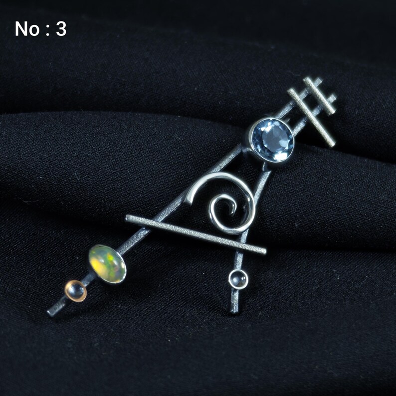 May include: A silver-toned brooch with an abstract design. It features a light blue gemstone, a swirling silver element, and an oval-shaped opal. The brooch has a geometric shape with the text "No: 3" in the upper left corner.