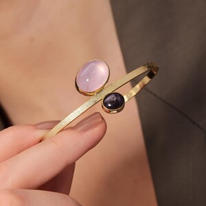 May include: Gold-toned bracelet featuring two gemstones: a large oval pink stone and a smaller, dark purple stone. The bracelet has a textured, matte finish and a minimalist design, suitable for everyday wear or special occasions.