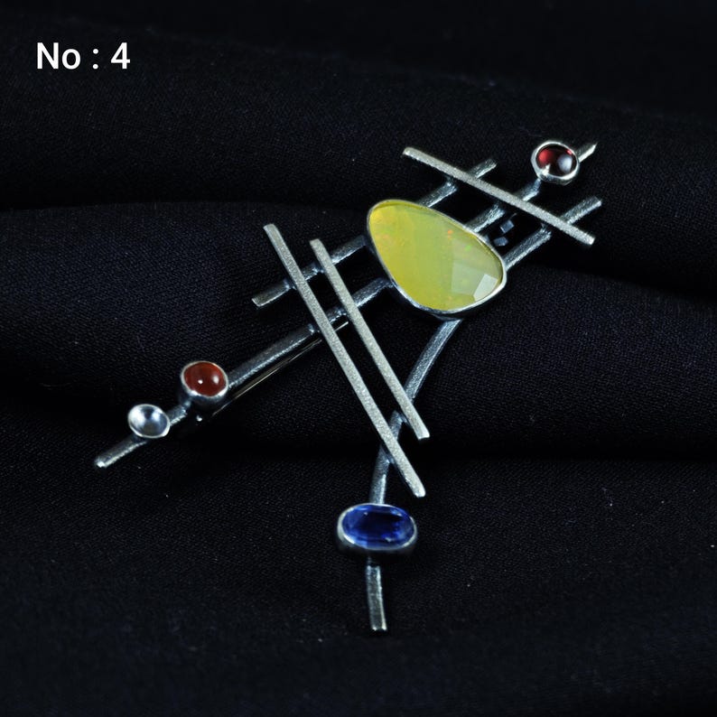 May include: Abstract silver-toned brooch with geometric design. Features a large, faceted yellow gemstone, a small blue gemstone, and two red gemstones. The brooch has a pin closure and is set against a black background.