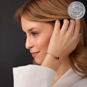 Könnte beinhalten: Ein goldfarbenes Armband mit zwei kleinen, runden Edelsteinen, einem blauen und einem roten. Das Armband hat ein zartes, minimalistisches Design. Das Bild enthält auch ein silbernes Emblem mit der Aufschrift "100% Handmade Sterling Silver Genuine Stone".