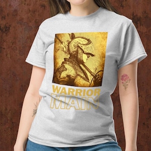 Peut inclure: T-shirt gris clair avec une illustration dorée d'un chevalier médiéval de profil, tenant une lance. Le texte "WARRIOR MAIN" est imprimé en or en dessous. Le t-shirt est fait d'une matière douce.