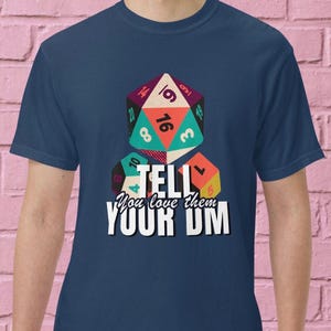 May include: Navy blue t-shirt featuring a colorful graphic of three dice, with the text "TELL You love them YOUR DM". The dice are in shades of orange, teal, and purple. The shirt is a short-sleeved crew neck.