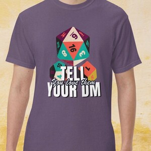 May include: A purple t-shirt with a graphic of dice and the text "TELL You love them YOUR DM". The dice are multi-coloured and have numbers on them. The text is in a bold, white font.
