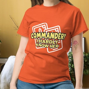 May include: An orange t-shirt with the text "COMMANDER? I HARDLY KNOW HER" in a white and yellow design. The graphic is set within two white card-shaped outlines. The shirt is a short-sleeved crew neck.